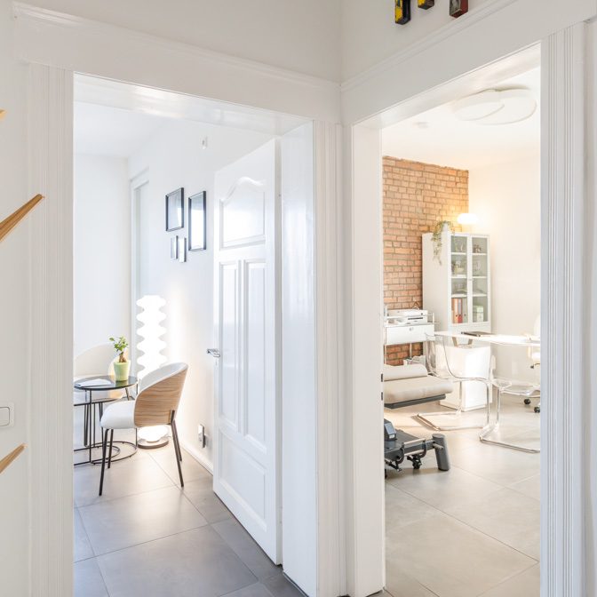 Heller Flur mit Blick auf einen Raum mit Therapieliege und Vitrinenschrank.