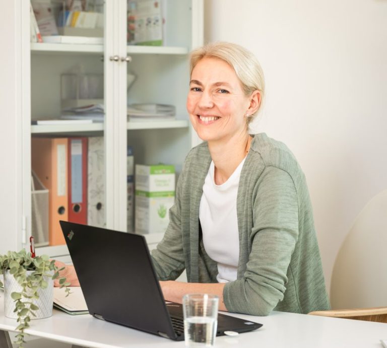 Lächelnde Frau mit Laptop am Tisch, Wasserglas und Pflanzen im Hintergrund.