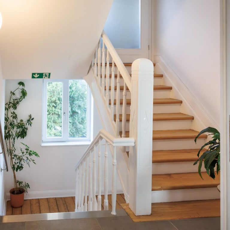 Treppenhaus mit Holzstufen, weißen Wänden und einer großen Fensterfront.