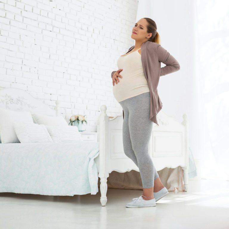 Schwangere Frau in einem hellen Raum, die sich an ihren Rücken hält.