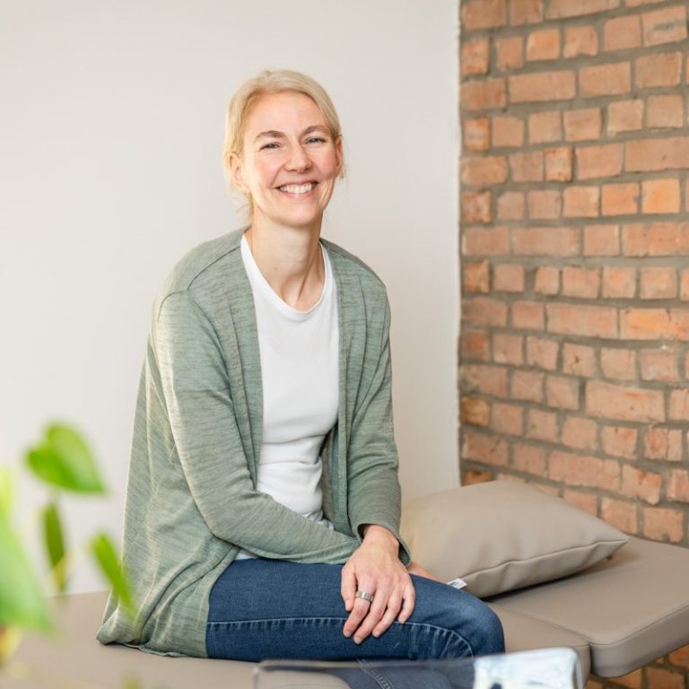 Lächelnde Frau mit blonden Haaren sitzt entspannt auf einer Therapieliege vor einer Backsteinwand.