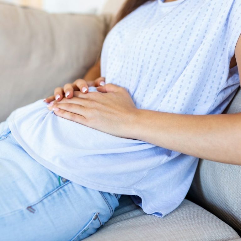 Frau sitzt auf einer Couch und hält ihren Bauch.