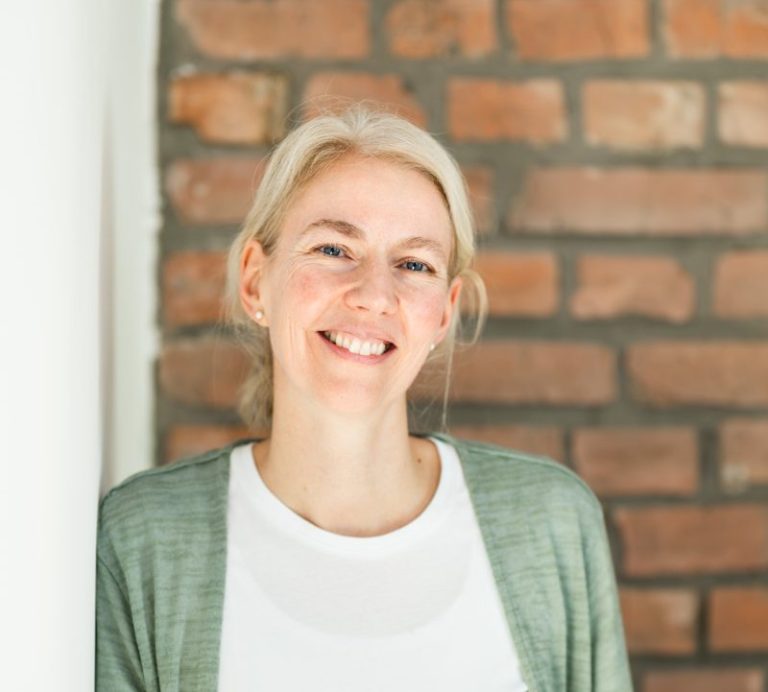 Lächelnde Frau mit blonden Haaren vor einer Backsteinwand.